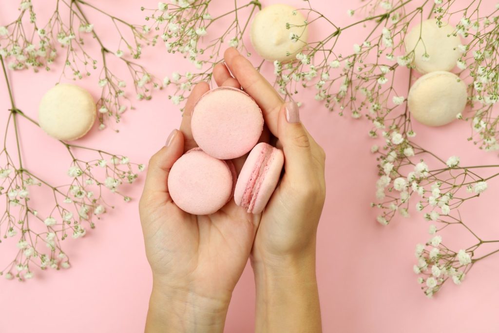 female hands hold macaroons on pink background with macaroons and flowers.jpg
